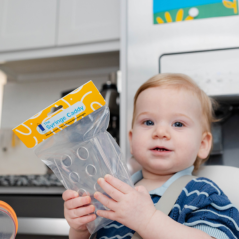 Baby in a high chair holding The Syringe Caddy, a medicine storage solution, in a kitchen setting.