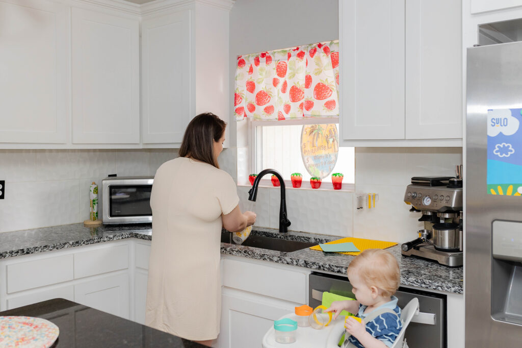 Mom washing syringes in a kitchen while baby plays at high chair